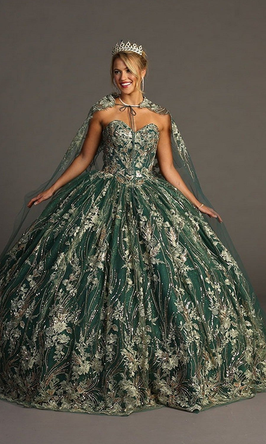 A woman poses with arms outstretched against a plain background, wearing the Quinceanera Dress CHF3079 By Chicasโa strapless green ballgown adorned with metallic floral embroidery and a jeweled crown.